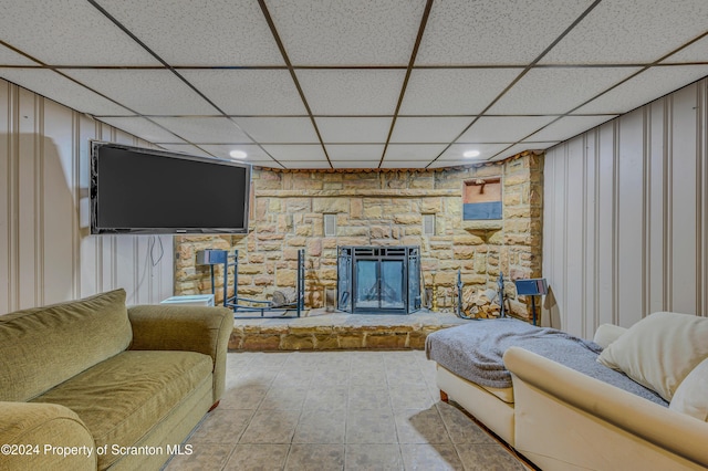 living room with a drop ceiling, light tile patterned flooring, a stone fireplace, and wooden walls