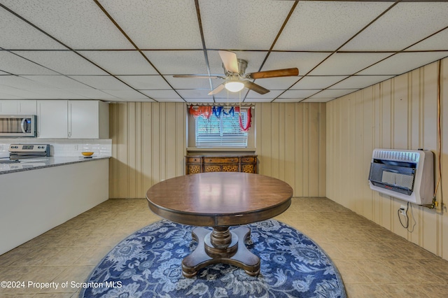 dining area with heating unit, a drop ceiling, wooden walls, and ceiling fan