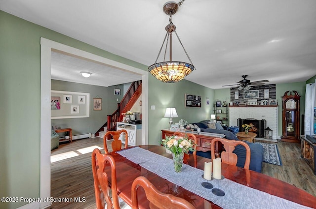 dining space with baseboard heating, ceiling fan, dark wood-type flooring, and a brick fireplace