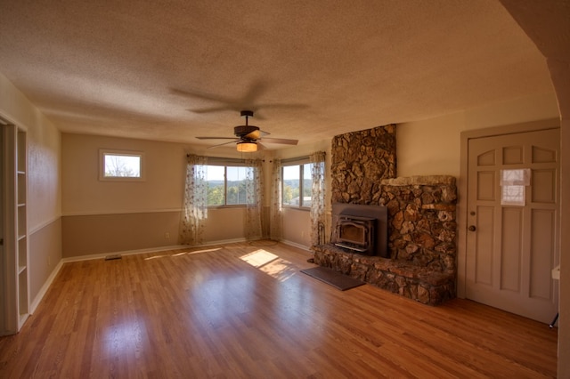 unfurnished living room featuring a stone fireplace, light hardwood / wood-style flooring, ceiling fan, a textured ceiling, and a wood stove