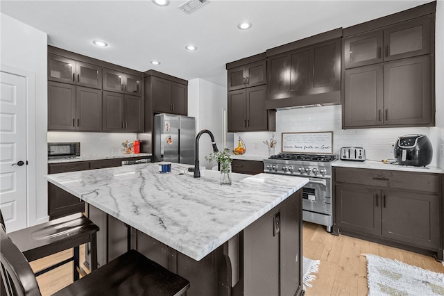 kitchen with light wood-type flooring, backsplash, an island with sink, appliances with stainless steel finishes, and a kitchen breakfast bar