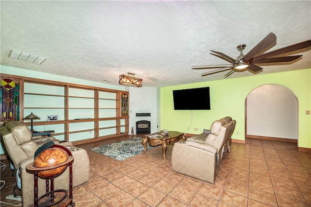 living room featuring ceiling fan, tile flooring, a brick fireplace, a textured ceiling, and brick wall