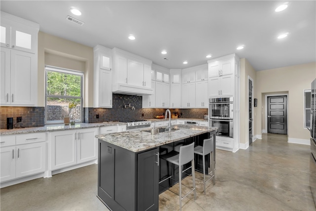 kitchen featuring backsplash, stainless steel double oven, white cabinets, and an island with sink