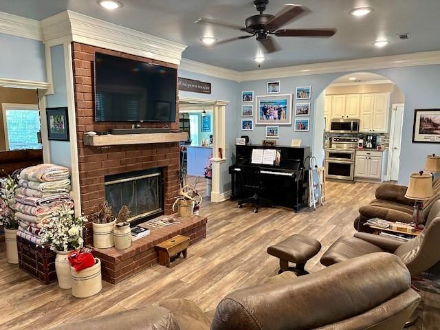 living room with a ceiling fan, light wood finished floors, a fireplace, arched walkways, and ornamental molding