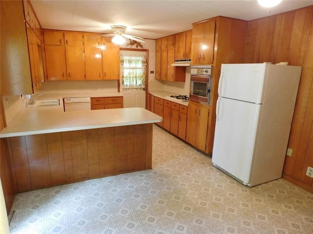 kitchen with kitchen peninsula, white appliances, ceiling fan, sink, and wood walls