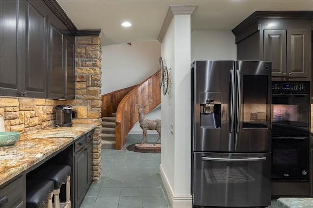 kitchen with wooden walls, stainless steel refrigerator with ice dispenser, black double oven, light tile patterned flooring, and dark brown cabinets