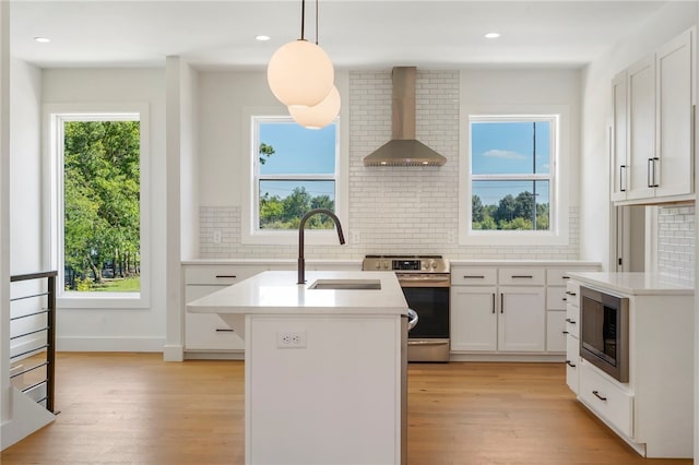 kitchen with white cabinets, wall chimney range hood, stainless steel appliances, and an island with sink