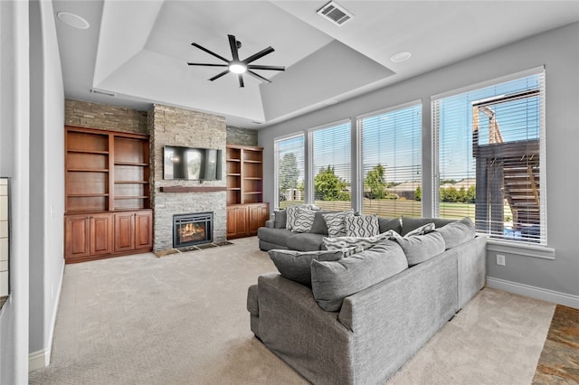 carpeted living room featuring a raised ceiling, a stone fireplace, ceiling fan, and built in shelves