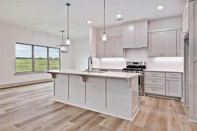 kitchen with sink, hanging light fixtures, stainless steel range with gas cooktop, an island with sink, and light wood-type flooring