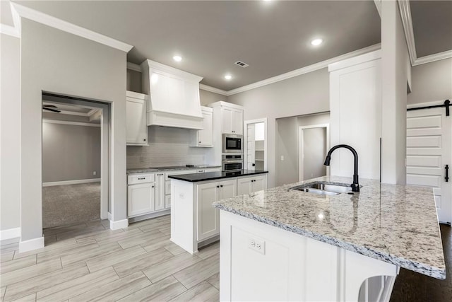 kitchen with a barn door, stainless steel appliances, a sink, custom exhaust hood, and an island with sink