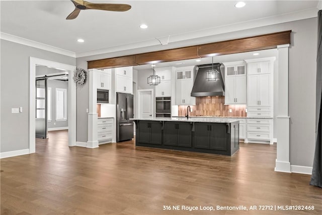 kitchen with custom exhaust hood, a kitchen island with sink, black appliances, a barn door, and white cabinetry