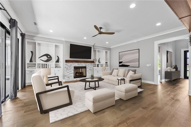 living room with built in shelves, ceiling fan, a stone fireplace, wood-type flooring, and ornamental molding