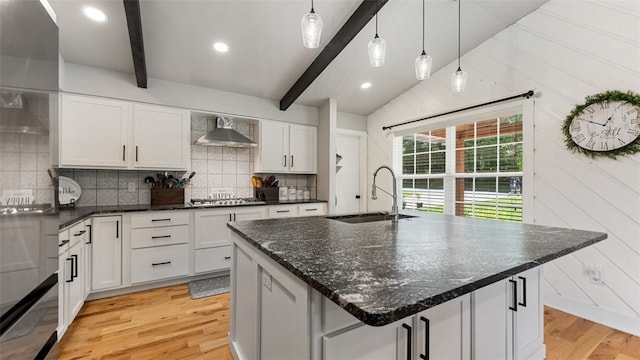 kitchen with lofted ceiling with beams, wall chimney exhaust hood, light wood-type flooring, and an island with sink