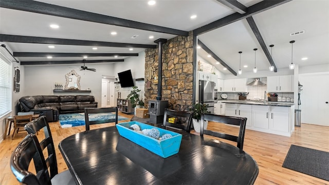 dining room with vaulted ceiling with beams, light hardwood / wood-style flooring, a wood stove, sink, and ceiling fan