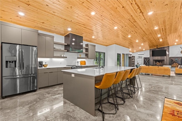 kitchen with a breakfast bar, gray cabinetry, a center island with sink, stainless steel fridge with ice dispenser, and wooden ceiling