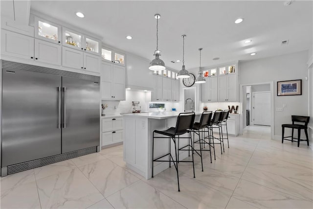 kitchen with built in fridge, a breakfast bar, white cabinets, hanging light fixtures, and a center island with sink