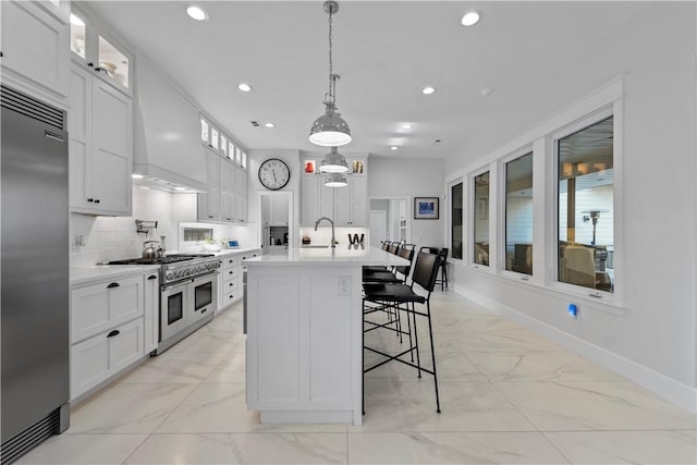 kitchen with pendant lighting, white cabinetry, high quality appliances, custom range hood, and a center island with sink