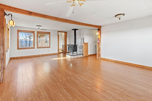 unfurnished living room with ceiling fan, beamed ceiling, a wood stove, a textured ceiling, and light hardwood / wood-style floors