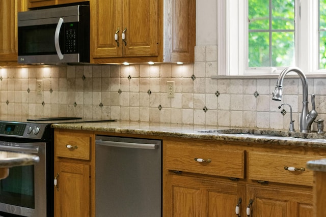 kitchen featuring stainless steel appliances, decorative backsplash, brown cabinetry, a sink, and light stone countertops