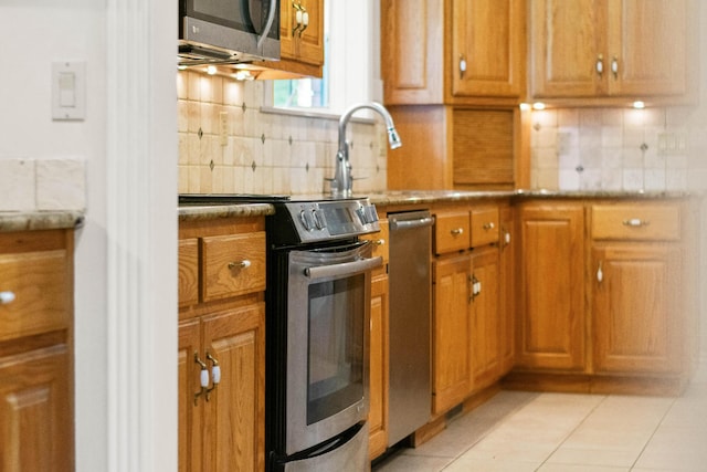 kitchen with light tile patterned floors, brown cabinetry, light stone countertops, stainless steel appliances, and backsplash