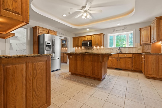 kitchen with stone counters, a center island, appliances with stainless steel finishes, brown cabinets, and a tray ceiling