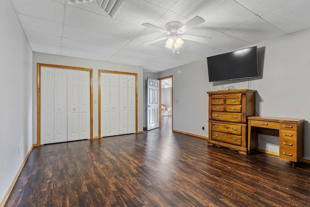 unfurnished bedroom with ceiling fan, dark hardwood / wood-style flooring, a drop ceiling, and two closets