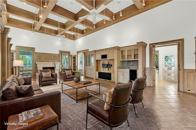 living room with a wainscoted wall, coffered ceiling, a high ceiling, a warm lit fireplace, and beamed ceiling