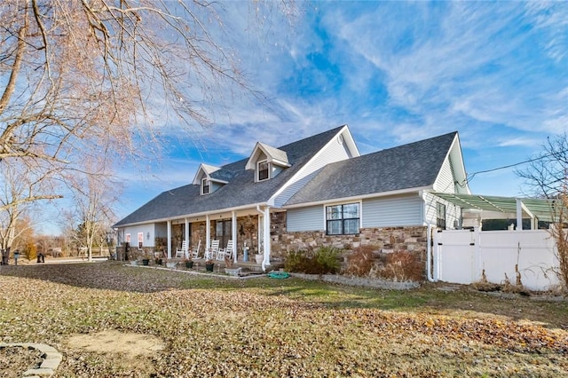 cape cod-style house with stone siding, fence, a porch, and roof with shingles