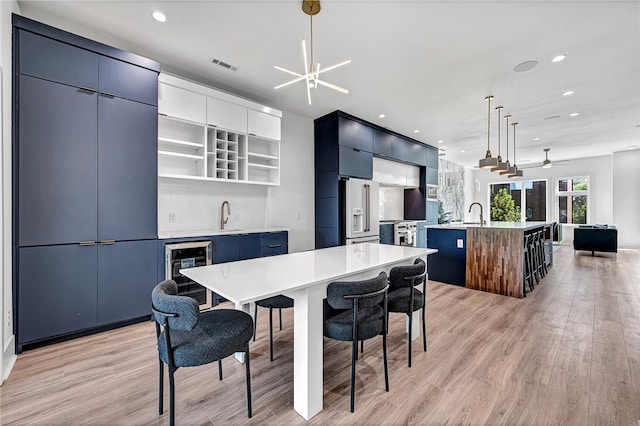 kitchen with decorative light fixtures, light wood-type flooring, ceiling fan with notable chandelier, an island with sink, and wine cooler