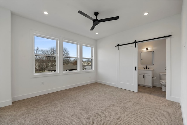 unfurnished bedroom featuring a barn door, ensuite bath, sink, ceiling fan, and light colored carpet