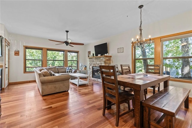 dining room with a stone fireplace, light wood-style flooring, ceiling fan with notable chandelier, and baseboards