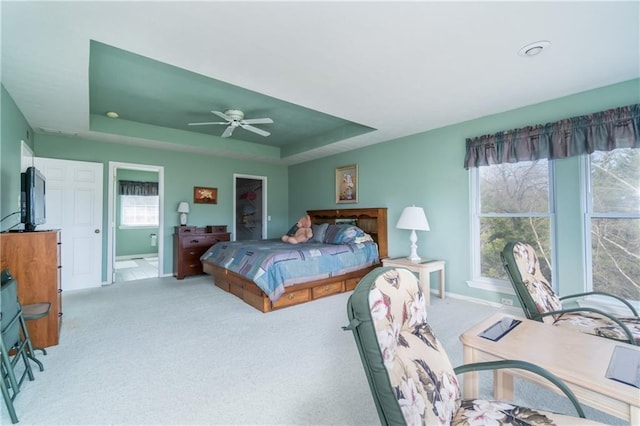 bedroom with ceiling fan, ensuite bathroom, a tray ceiling, light carpet, and a walk in closet