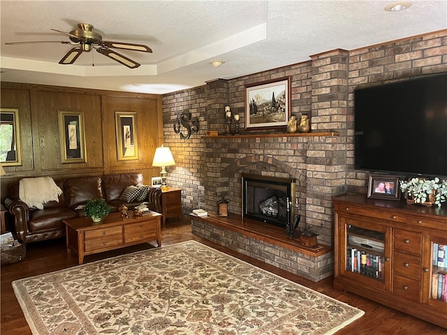 living room featuring a textured ceiling, a brick fireplace, a raised ceiling, dark hardwood / wood-style flooring, and ceiling fan