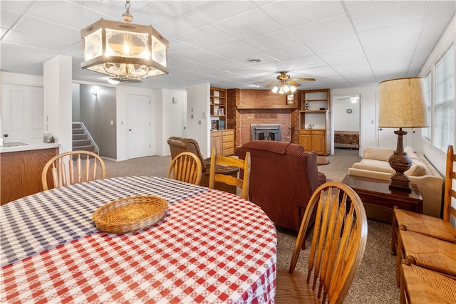 carpeted dining space featuring built in shelves, a fireplace, a drop ceiling, and ceiling fan with notable chandelier