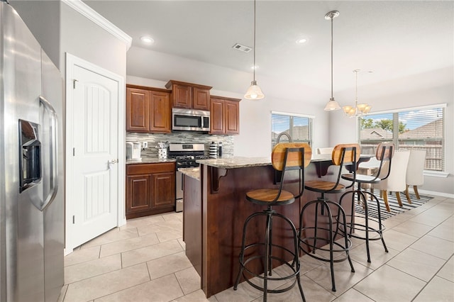 kitchen with a kitchen island with sink, hanging light fixtures, light stone countertops, appliances with stainless steel finishes, and tasteful backsplash