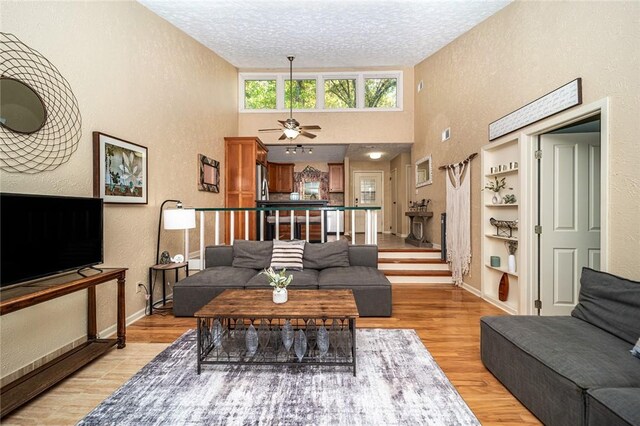 living room with built in shelves, ceiling fan, a towering ceiling, a textured ceiling, and light wood-type flooring