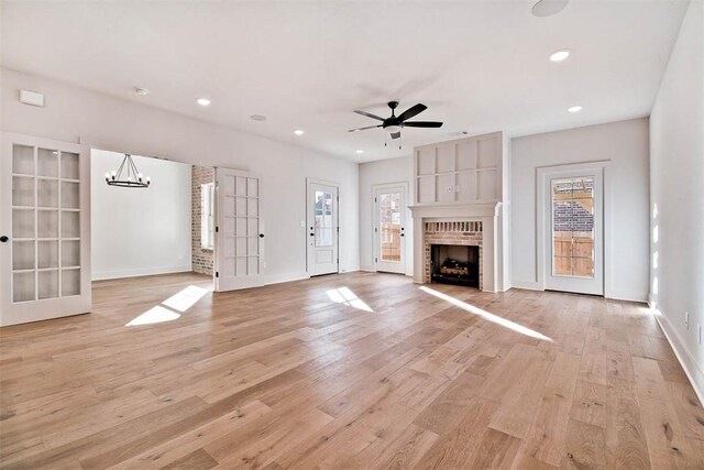 unfurnished living room with french doors, a brick fireplace, ceiling fan with notable chandelier, and light hardwood / wood-style flooring