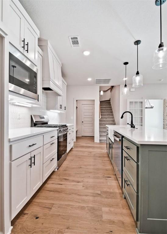 kitchen featuring appliances with stainless steel finishes, white cabinetry, hanging light fixtures, a center island with sink, and light wood-type flooring