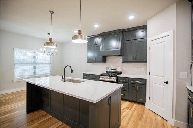 kitchen featuring sink, stainless steel gas range oven, an island with sink, decorative light fixtures, and custom range hood