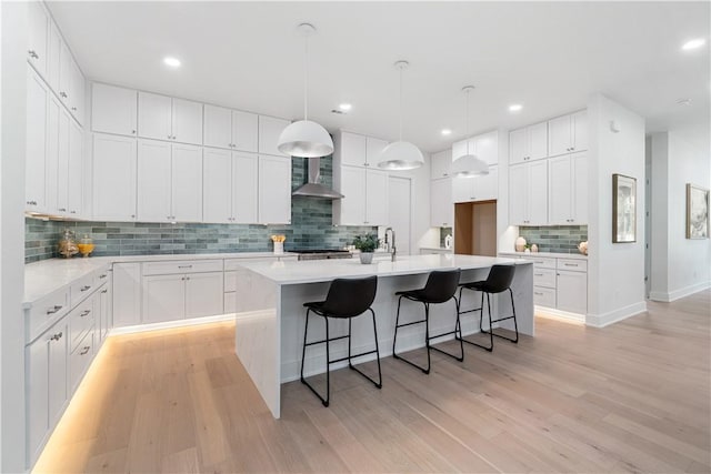 kitchen featuring light wood-type flooring, wall chimney exhaust hood, a kitchen island with sink, a kitchen bar, and tasteful backsplash