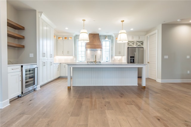 kitchen with custom range hood, white cabinetry, wine cooler, pendant lighting, and a kitchen island with sink