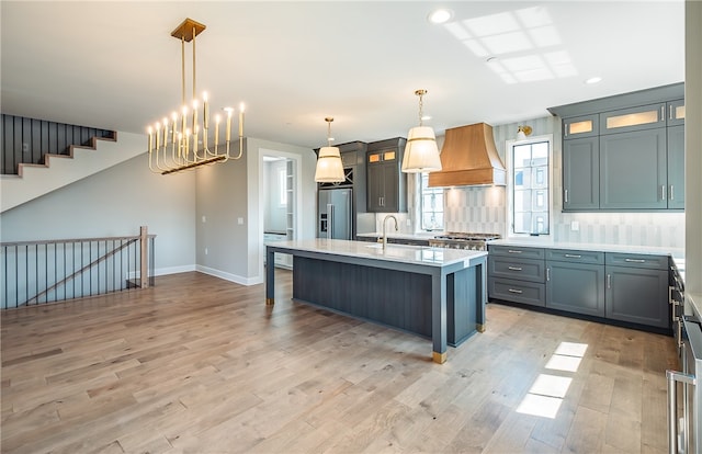 kitchen with pendant lighting, premium range hood, an island with sink, high end fridge, and light wood-type flooring