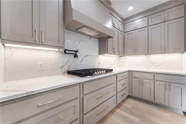 kitchen with light stone countertops, wall chimney range hood, stainless steel gas cooktop, backsplash, and light wood-type flooring