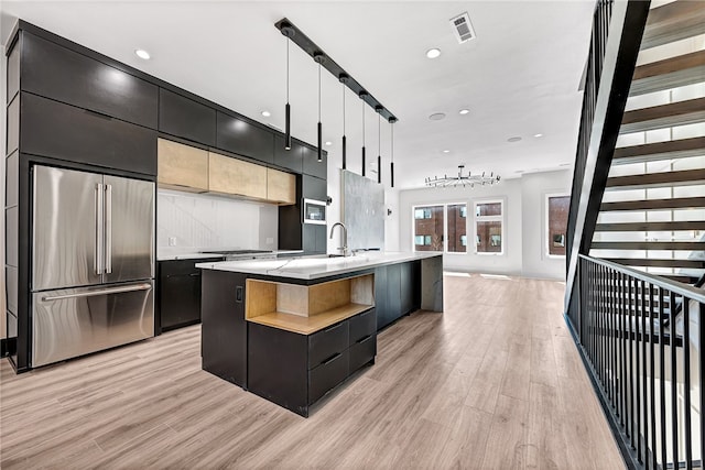 kitchen with an island with sink, light wood-type flooring, sink, hanging light fixtures, and high quality fridge