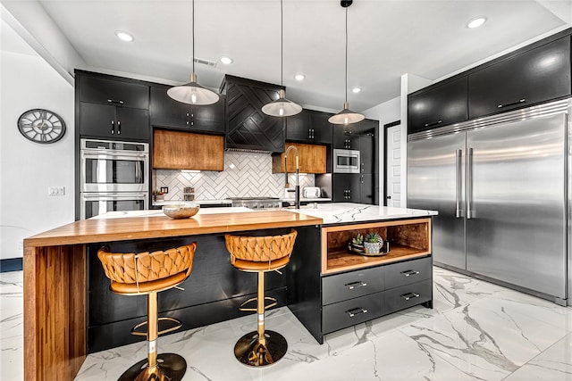 kitchen featuring an island with sink, tasteful backsplash, premium range hood, built in appliances, and decorative light fixtures