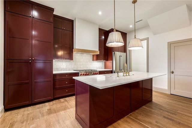 kitchen with an island with sink, appliances with stainless steel finishes, decorative light fixtures, light wood-type flooring, and custom range hood