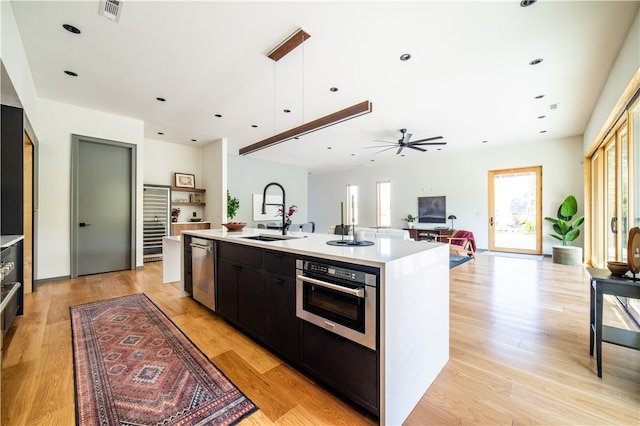 kitchen with appliances with stainless steel finishes, ceiling fan, sink, a center island with sink, and light hardwood / wood-style flooring