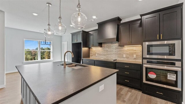 kitchen with premium range hood, a center island with sink, stainless steel oven, decorative light fixtures, and sink