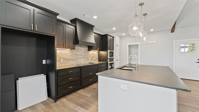 kitchen featuring wall chimney range hood, sink, hanging light fixtures, a kitchen island with sink, and stainless steel oven