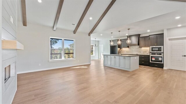 kitchen with an island with sink, stainless steel appliances, decorative backsplash, decorative light fixtures, and beamed ceiling